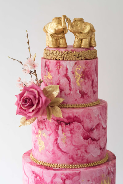 For a fusion wedding in Cheshire, the   wedding cake is hand-painted in Magenta to create a marbled effect and has gold details including two gold elephants stood on top of the cake.