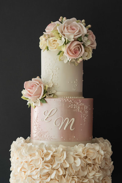 Effloresence wedding cake design. Pretty three-tiered wedding cake in ivory and shimmering pink with ivory ruffles, pale pink roses and delicate lace detail. Yorkshire wedding cake