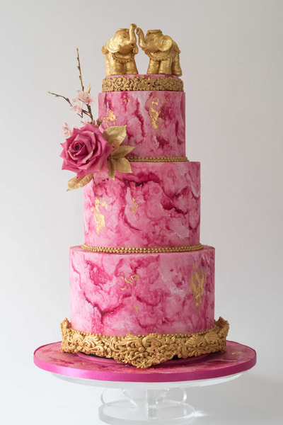 For a fusion wedding in Cheshire, the   wedding cake is hand-painted in Magenta to create a marbled effect and has gold details including two gold elephants stood on top of the cake.