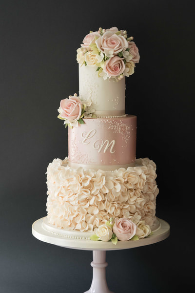Effloresence wedding cake design.  Pretty three-tiered wedding cake in ivory and shimmering pink with ivory ruffles, pale pink roses and delicate lace detail