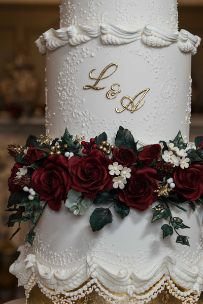 The bottom tier of this wedding cake is separated from the top two tiers by a ring of deep red sugar flowers, gold berries and handmade pine-cones